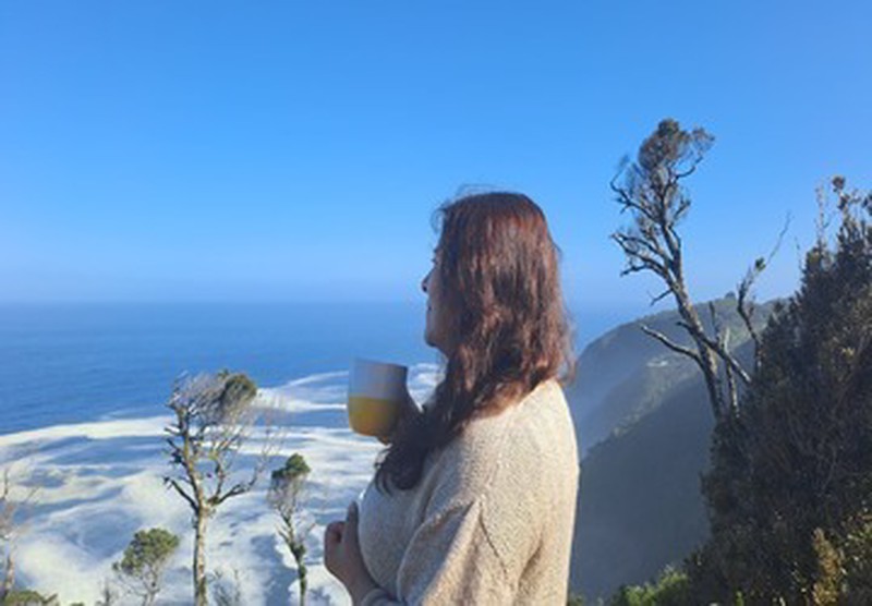 Mujer de perfil contemplando bosque y océano en Ko Glamping mientras sostiene taza.