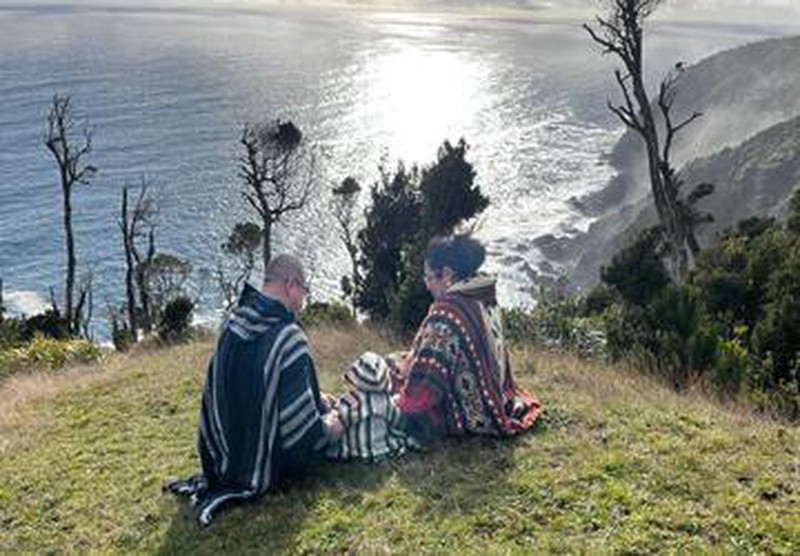 Familia con ponchos, dos adultos y bebé, de espaldas, contemplando mar y árboles en Ko Glamping.